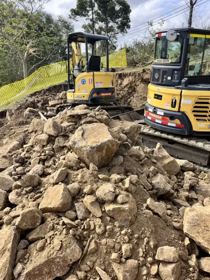 An excavator working through broken rock and boulders on a cleared, benched block.