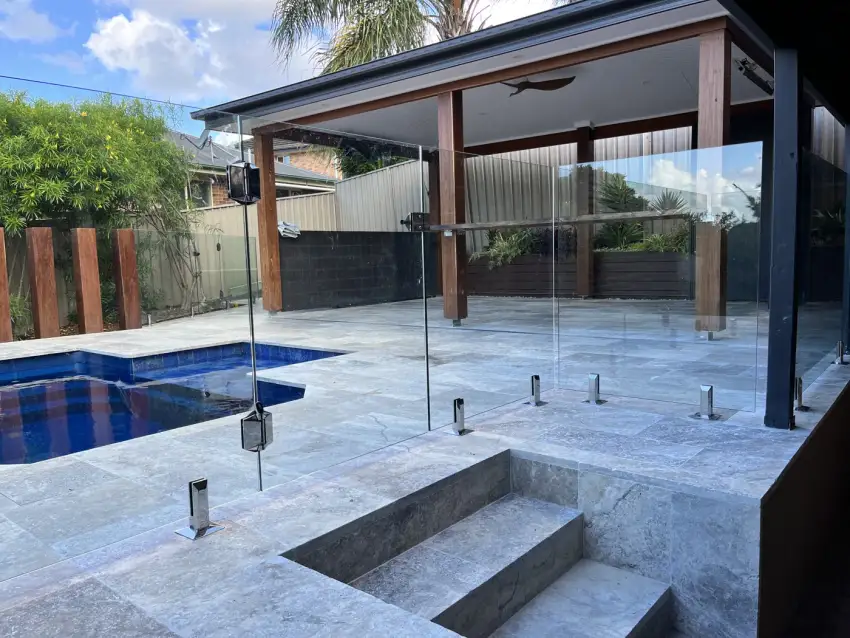 A travertine-paved pool and spa surround under a covered timber-post alfresco, with frameless glass fencing.