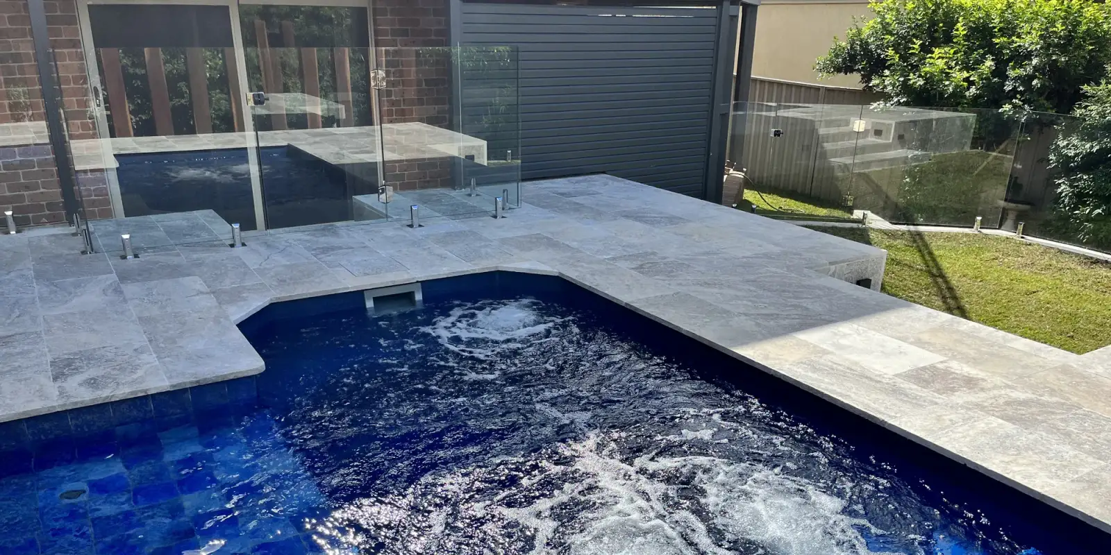 A finished Lattas Landscaping pool and spa in Shellharbour: a deep-blue tiled pool with bubbling spa jets, travertine paving and frameless glass fencing.