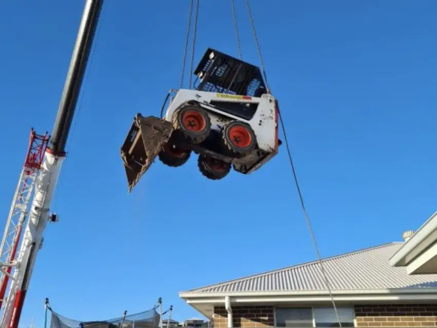 A crane lifting a skid-steer loader over a fence into a tight-access backyard.