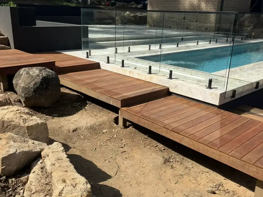 A frameless glass pool fence with a timber step-deck crossing sandstone boulders.