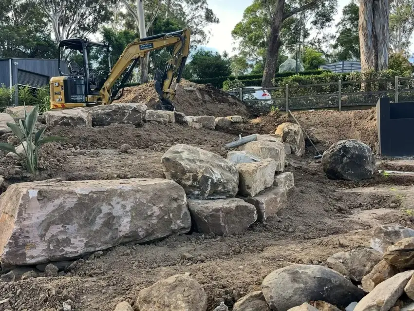 An excavator setting large sandstone boulders into a retaining wall on a cleared Illawarra site.