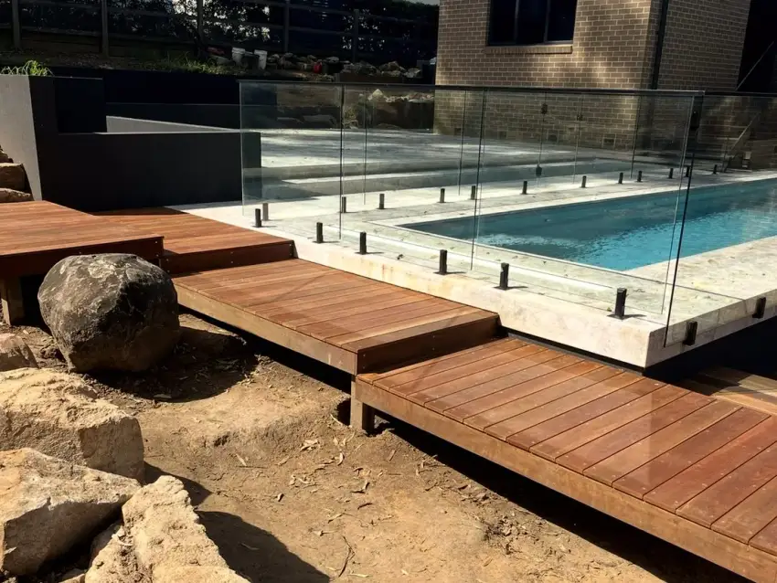 A frameless glass pool fence beside a timber deck bridge over sandstone boulders, with a brick home behind.