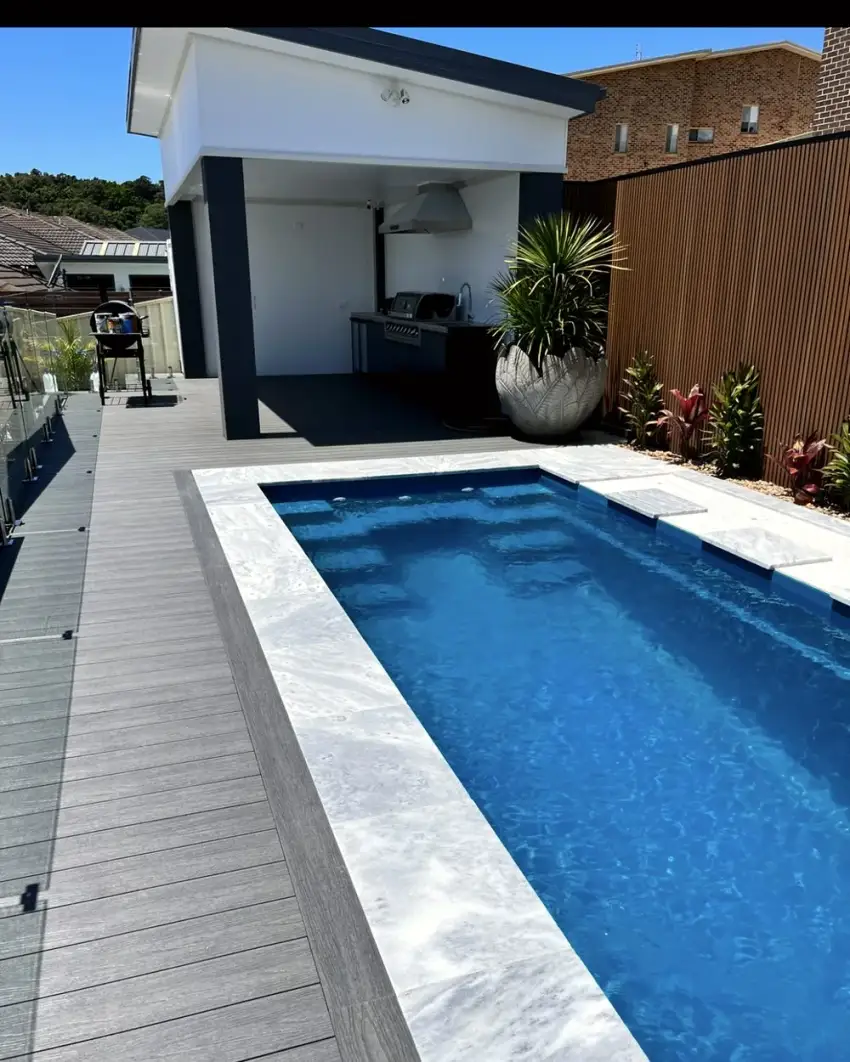 A finished concrete pool with pale stone coping, timber decking and a covered alfresco kitchen built by Lattas Landscaping.